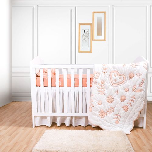 A crib with a white fitted sheet and a quilt with floral and heart embroidery in pink and white colors is displayed in the crib.