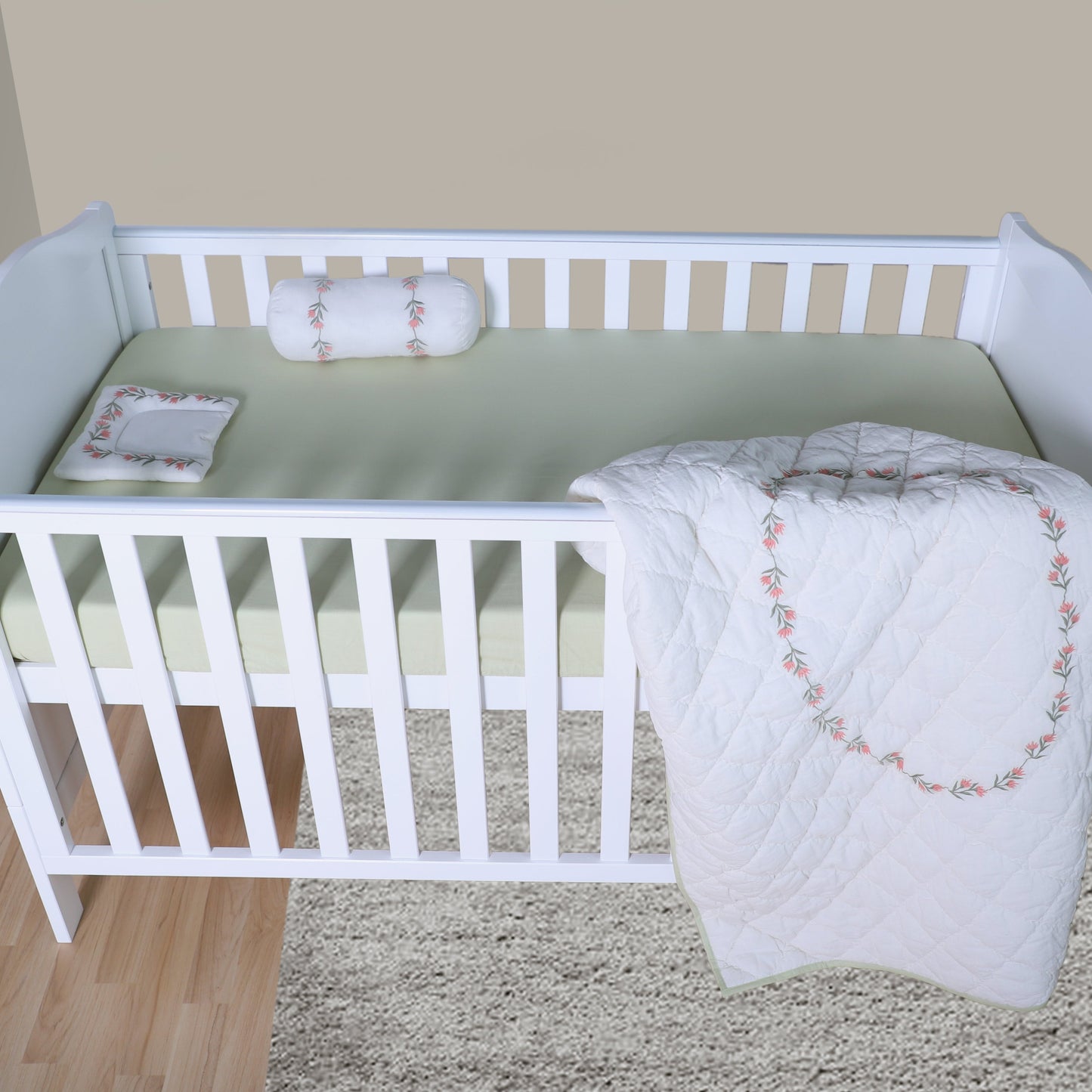 White crib with green mattress and white blanket with pink embroidery on a beige carpet.