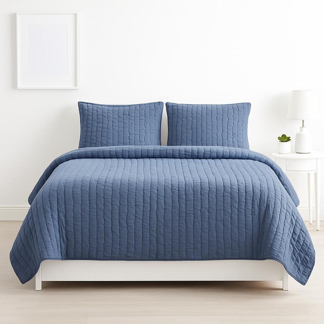 Blue quilted coverlet set on a bed in a room with a white wall and small plant.