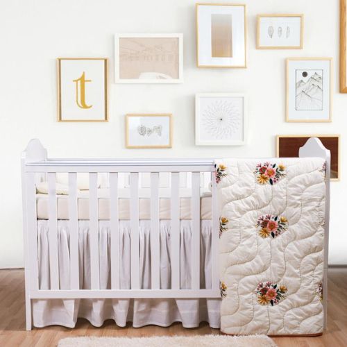 A baby quilt with white fabric and embroidered flowers, displayed next to a crib, with a room full of decor in the background.