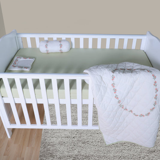 White crib with green mattress and white blanket with pink embroidery on a beige carpet.