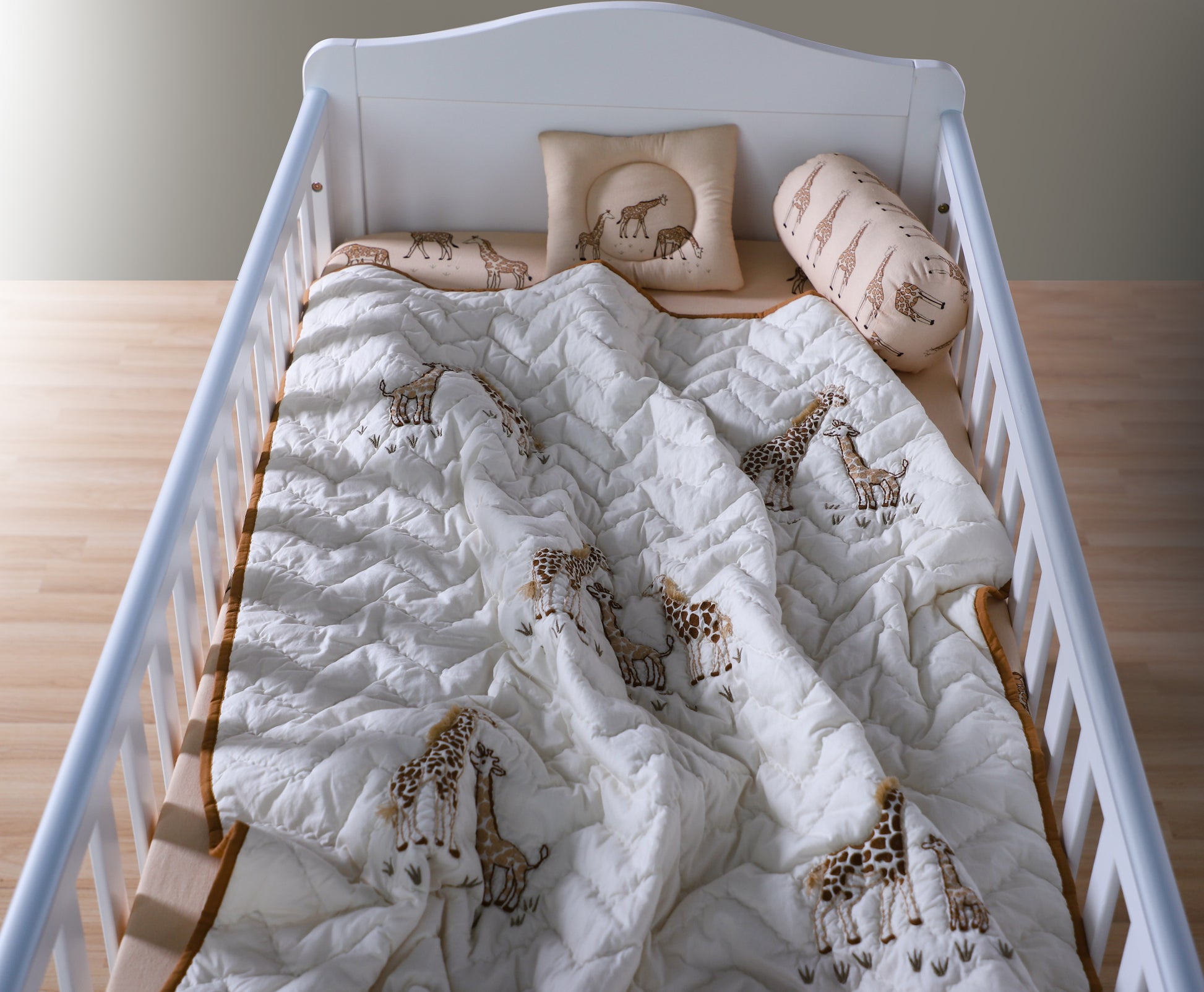 Crib with white quilt featuring giraffe patterns on a wooden floor.