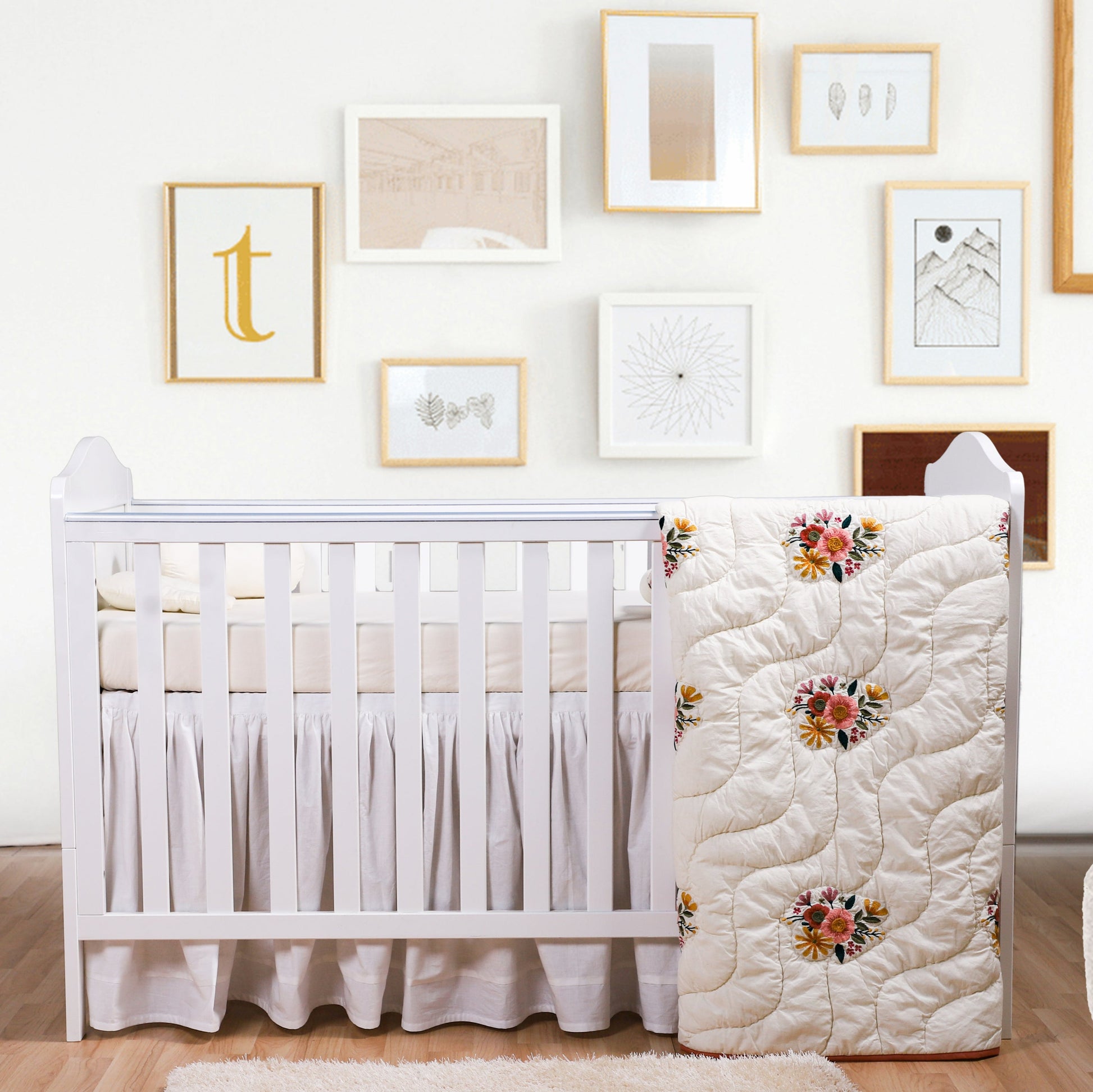 Nursery room with a white crib and floral quilt against a wall with framed pictures.