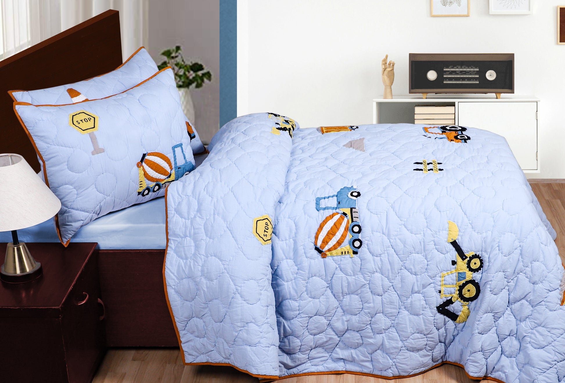 Children's bed with blue quilt featuring cartoon vehicles, nightstand with lamp, and decorative items on shelf.