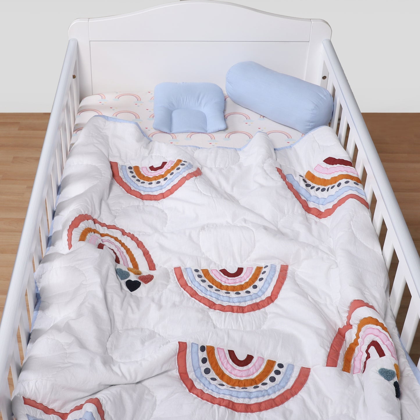 Crib with rainbow-patterned bedding and blue pillows on a wooden floor.