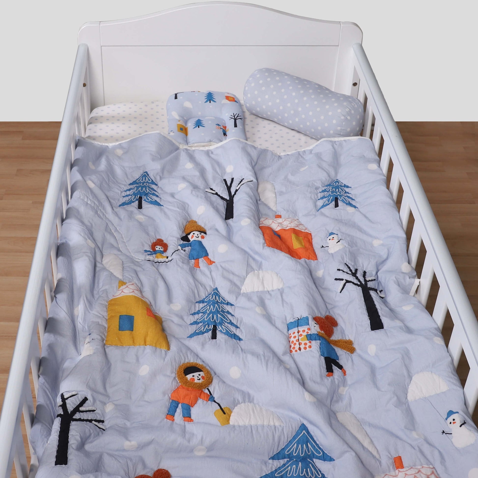 A baby quilt with a winter theme, featuring characters such as a snowman, trees, and possibly a bear, is displayed on a crib.