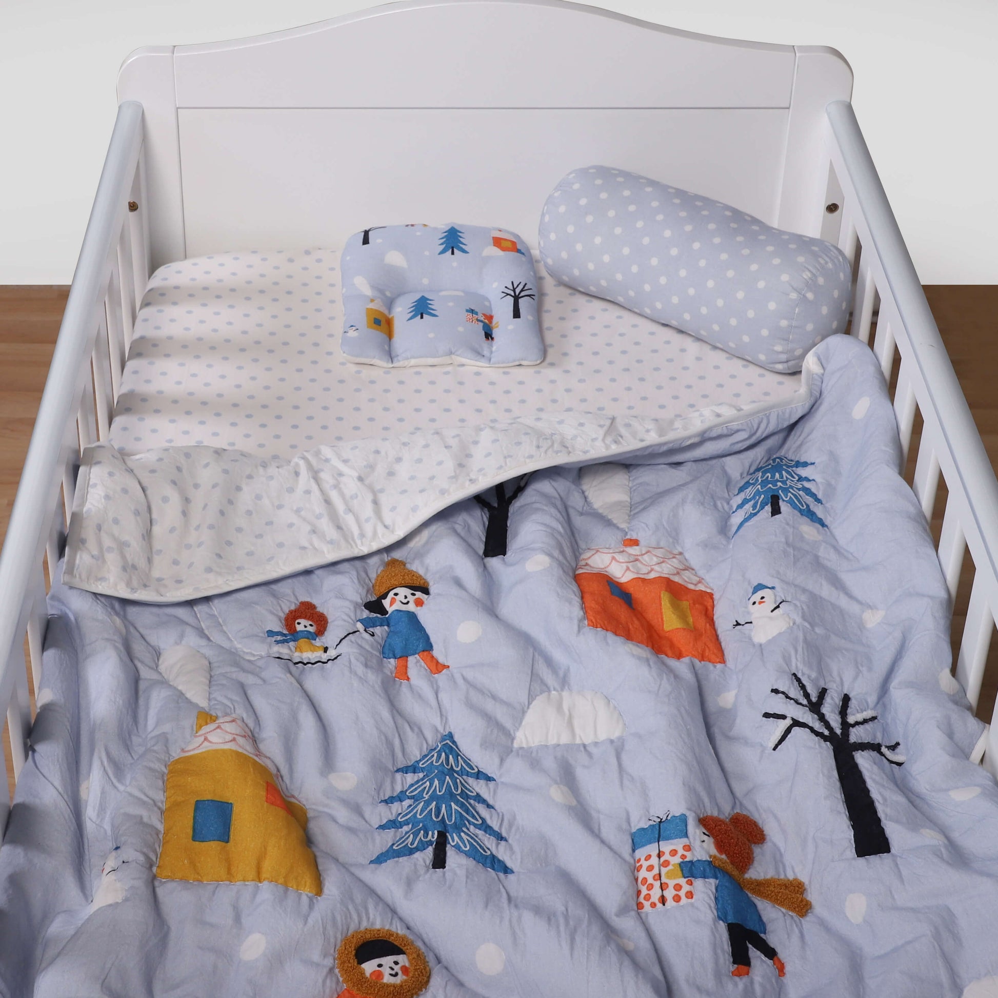 A baby quilt with a winter theme, featuring characters such as a snowman, trees, and possibly a bear, is displayed on a crib.