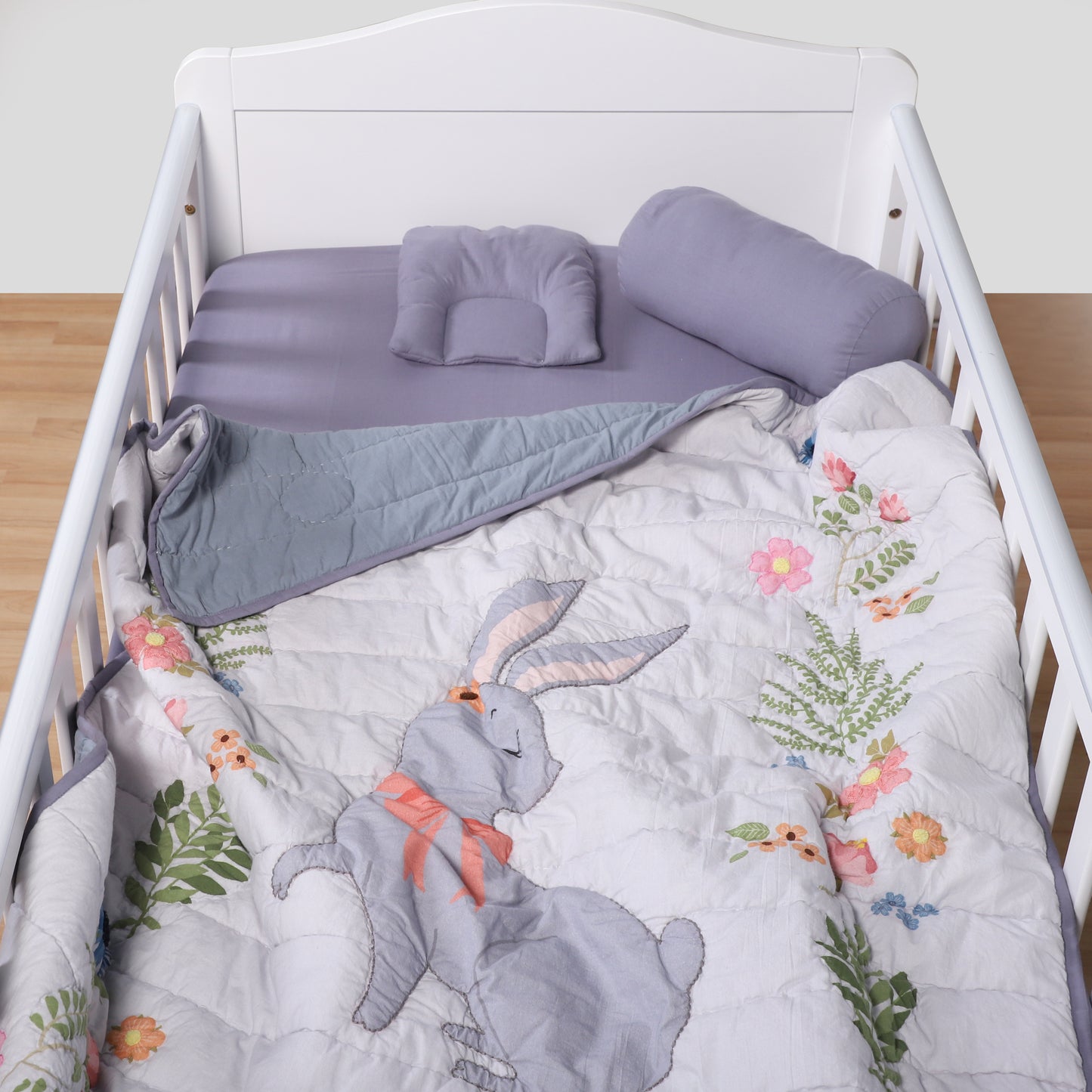 Crib with floral and rabbit design bedding, gray blanket, and purple pillows.