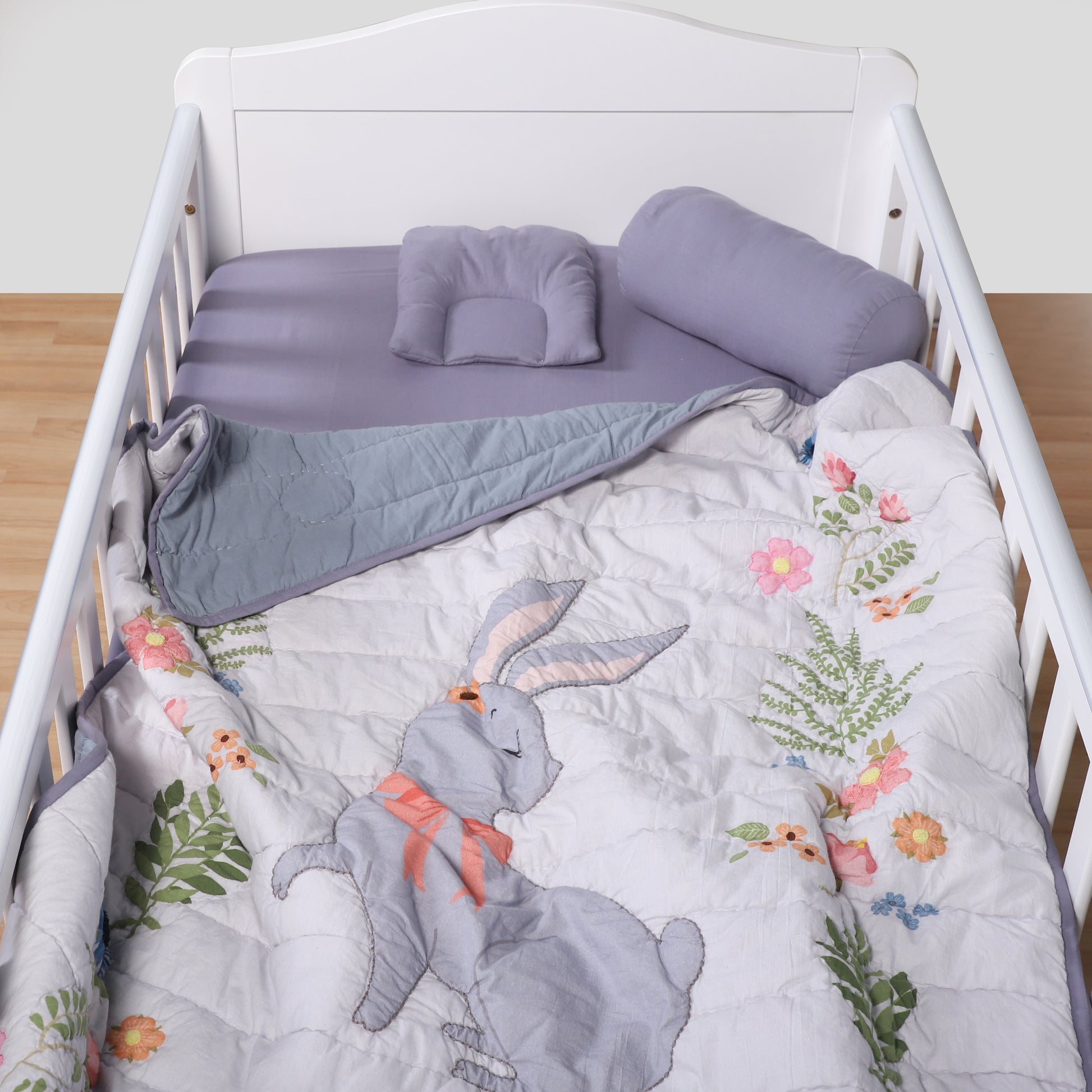 Crib with floral and rabbit design bedding, gray blanket, and purple pillows.
