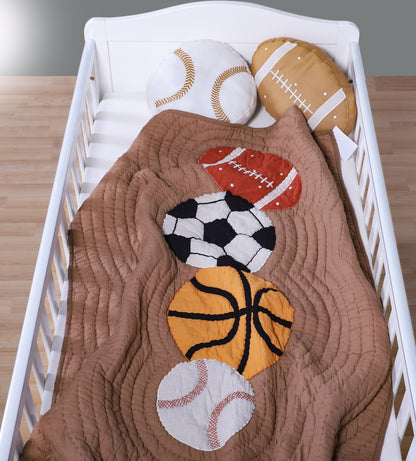 Crib with a brown quilt featuring sports ball appliqués on a wooden floor.