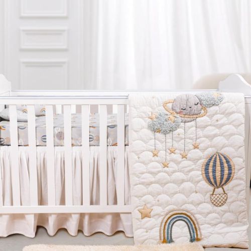 A baby crib set with a quilt featuring rainbow and hot air balloon designs, a fitted sheet, and a baby pillow, all in a matching soft color palette.