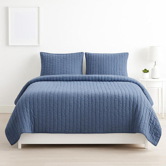 Blue quilted coverlet set on a bed in a room with a white wall and small plant.