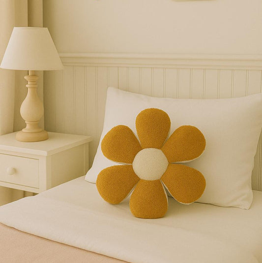 Yellow flower-shaped pillow on a bed with a lamp and decorative wall items in the background.
