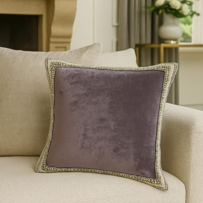 Purple velvet pillow with gold trim on a beige sofa in a living room setting.