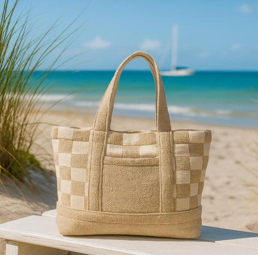 Beige checkered tote bag on a wooden bench with a beach and sailboat in the background