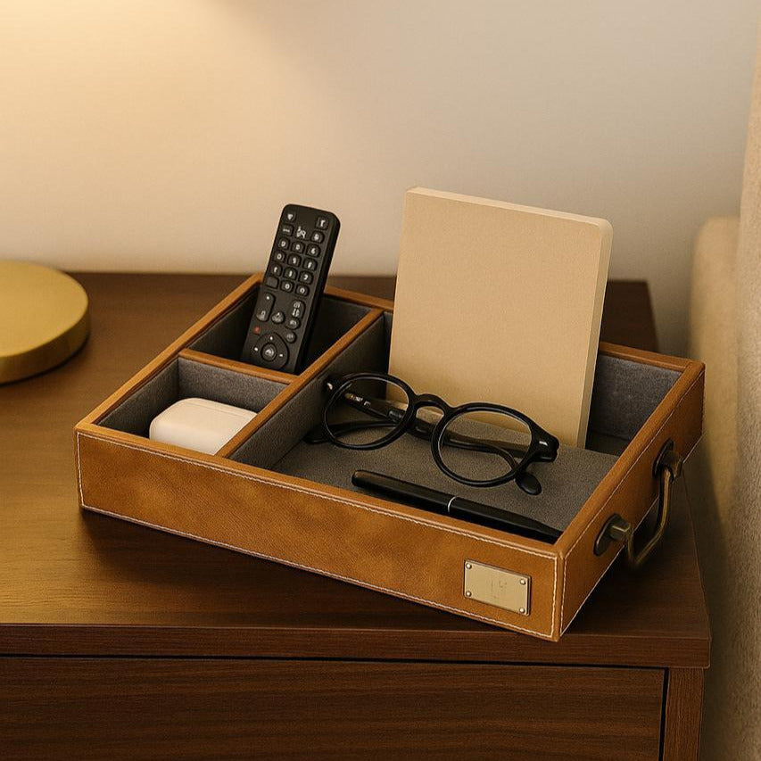 Brown leather tray with compartments holding a remote control, glasses, and a pen on a wooden surface.